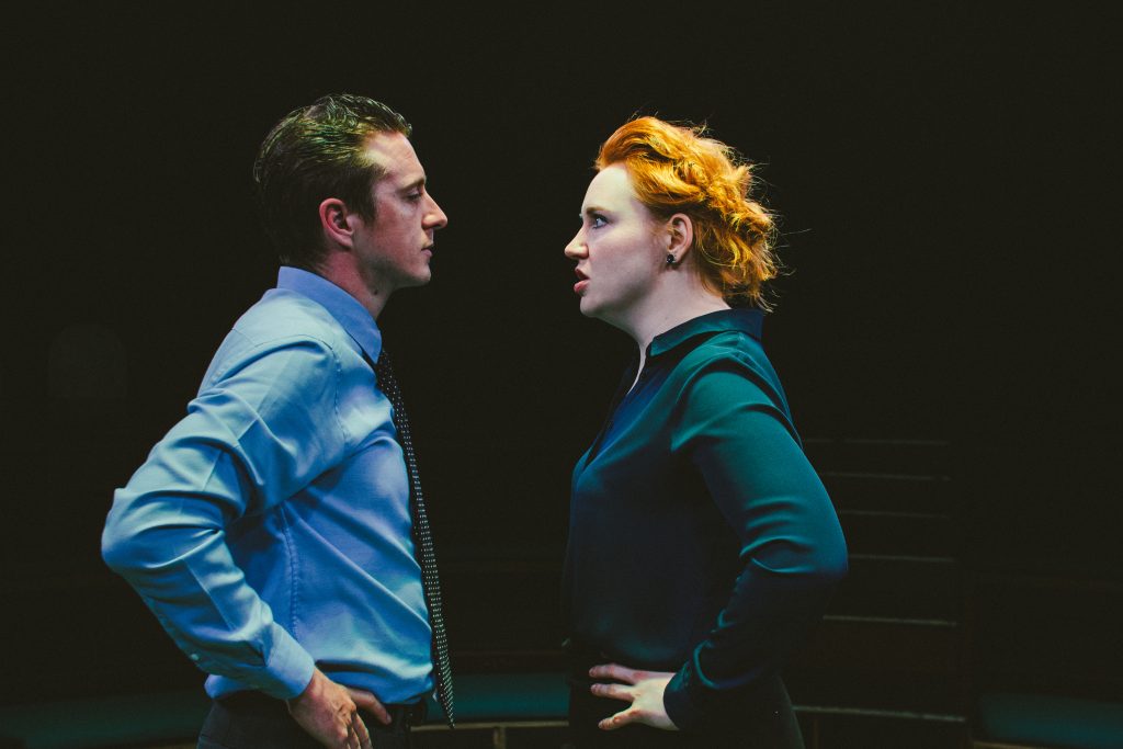 A man in a dress shirt and tie and a woman in a dark blouse stand facing each other on a stage.