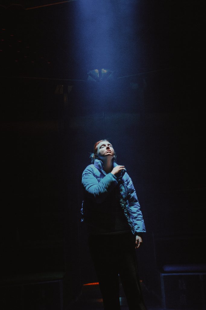 A person in a puffer jacket stands in a dark room, illuminated by a single spotlight from above.