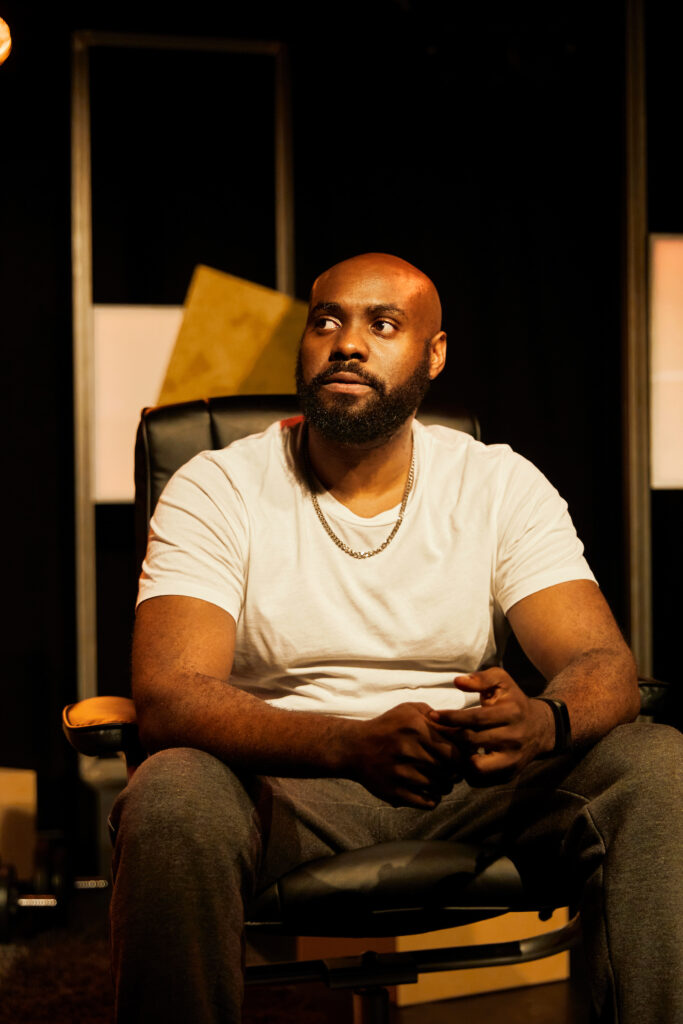 Actor Joseph Black as Tunde in Sessions. Tunde wears a white t-shirt and grey tracksuit bottoms, he sits in his flat looking around.