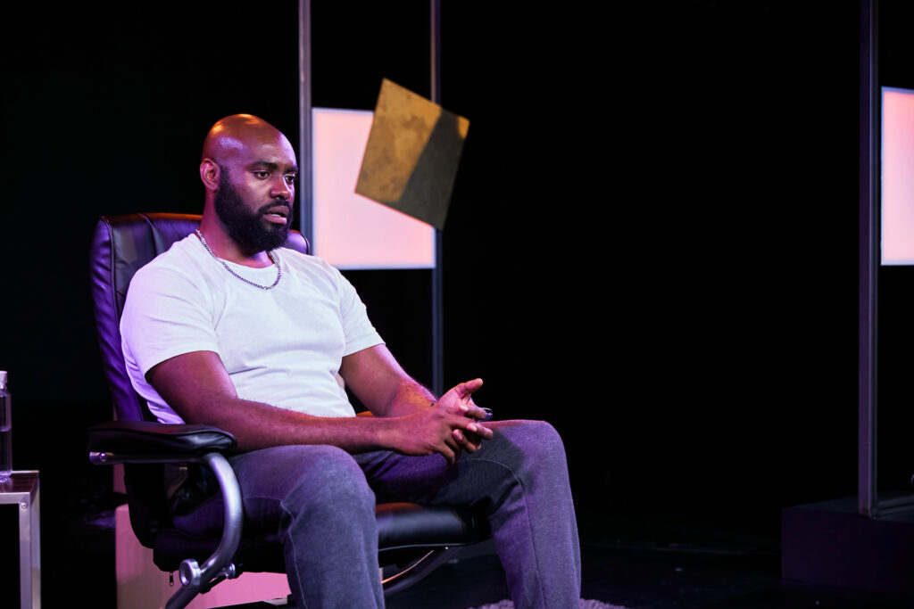 Actor Joseph Black as Tunde in Sessions. Tunde sits in an office chair wearing a white t-shirt and grey tracksuit bottom, his hands and interwoven as he sits.