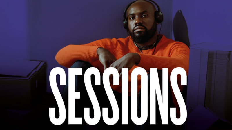 Actor Joseph Black sits on the floor with is arms across his legs, he wears headphones and an orange jumper as he looks directly into the camera. The typeface Sessions is in the foreground of the image.