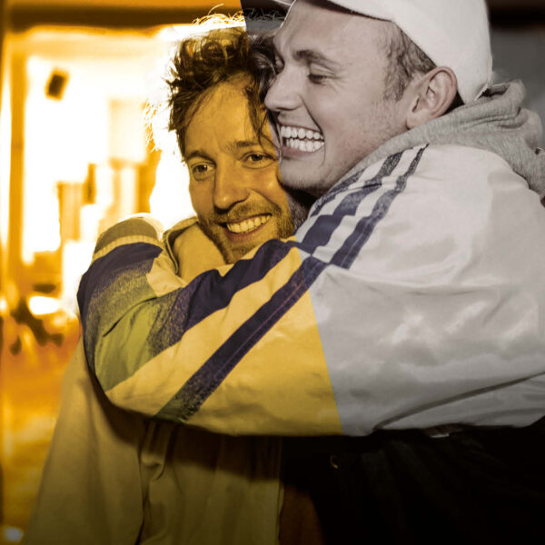 2 men stand hugging in a bar, both have huge smiles. One man wears a baseball cap and tracksuit top, the other man wears a casual jacket.