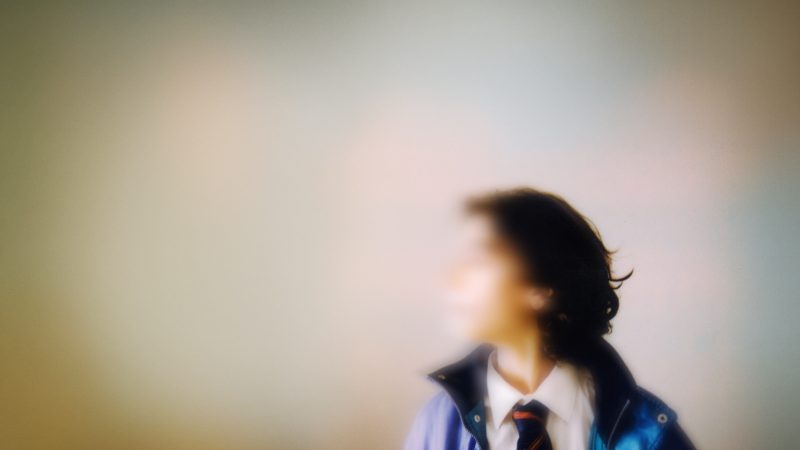 obscured and blurred image of a young person with dark hair wearing a white shirt and dark tie, with a blue anorak overcoat